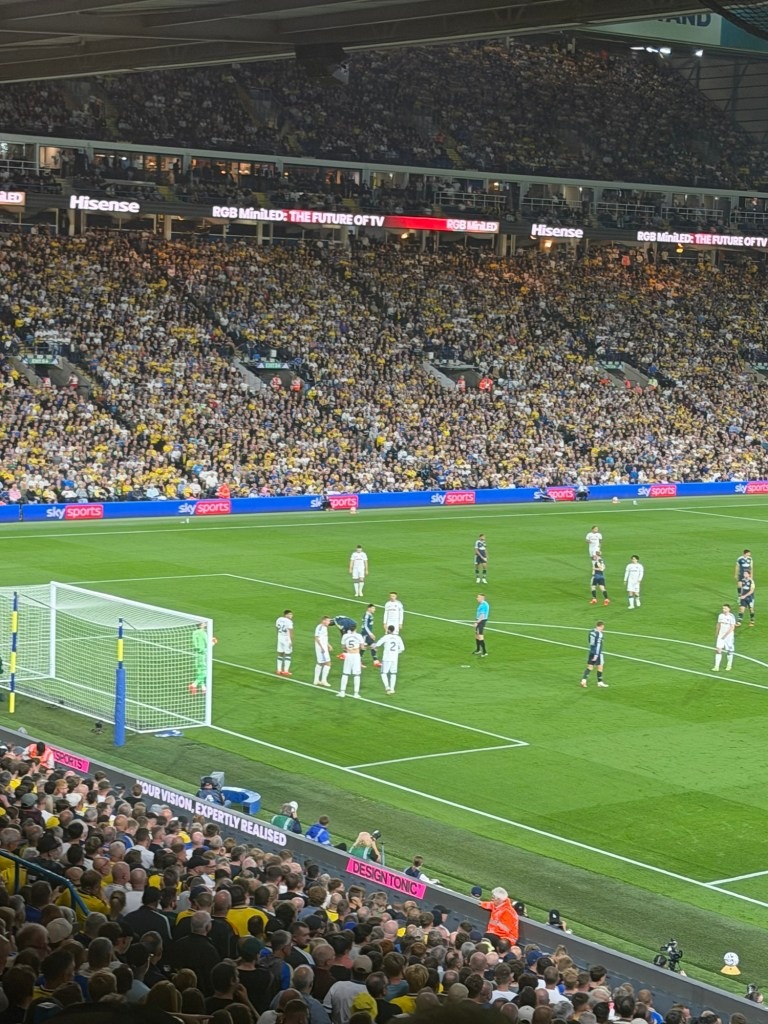 A crowded football stadium with fans in yellow and white, watching a match. Players are positioned around the penalty area, preparing for a set piece, while a referee oversees the play.